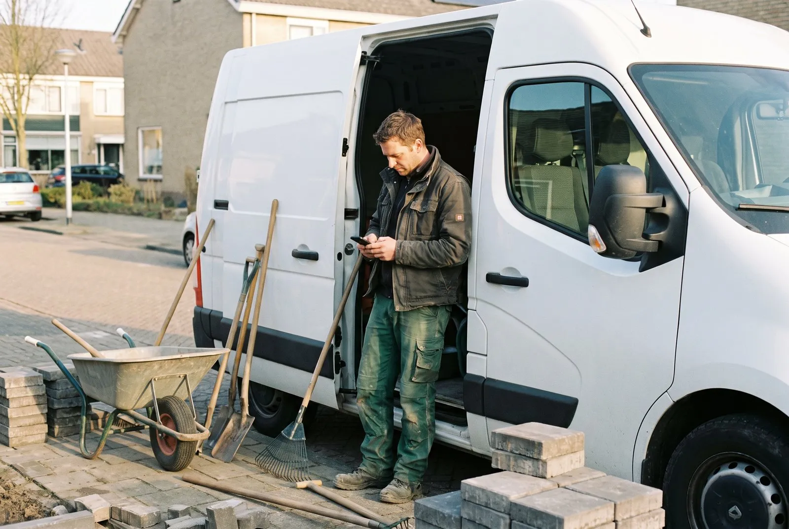 Een hovenier staat bij de open schuifdeur van zijn bus en kijkt op zijn telefoon, met een kruiwagen en gereedschap naast zich.
